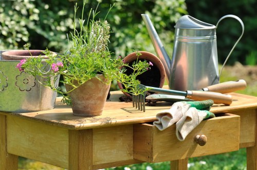 In the outdoor garden of a residential property in Beckenham, a wooden garden table is arranged with gardening tools and plant containers. The table holds a metal watering can, two terracotta pots, one filled with a variety of green herbs and flowering plants, and another empty pot. A pair of gardening gloves, a hand rake, and a small fork are also placed on the table, suggesting ongoing plant care activities. The background features lush green foliage, indicating a well-maintained garden setting. The scene is illuminated by natural daylight, creating a bright and inviting atmosphere appropriate for gardening and lawn care services in the Beckenham area, within postcode BR3 or nearby locales.